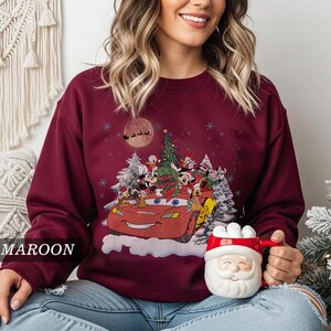 K&ouml;nnte beinhalten: Burgunderfarbenes Sweatshirt mit einem festlichen Aufdruck eines roten Autos voller Disney-Figuren, einem Weihnachtsbaum und Schneeflocken. Das Design umfasst Micky Maus, Donald Duck und andere Charaktere. Der Text "Merry Christmas" ist sichtbar.