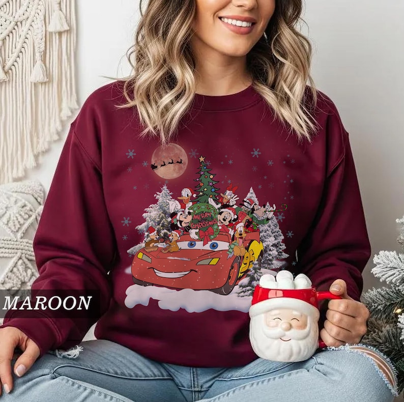 K&ouml;nnte beinhalten: Burgunderfarbenes Sweatshirt mit einem festlichen Aufdruck eines roten Autos voller Disney-Figuren, einem Weihnachtsbaum und Schneeflocken. Das Design umfasst Micky Maus, Donald Duck und andere Charaktere. Der Text "Merry Christmas" ist sichtbar.