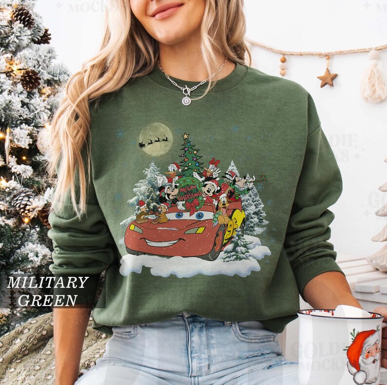 K&ouml;nnte beinhalten: Milit&auml;rgr&uuml;nes Sweatshirt mit einem weihnachtlichen Design. Es zeigt ein rotes Cartoon-Auto voller Disney-Figuren, einen Weihnachtsbaum und den Schriftzug "Merry Christmas". Das Design umfasst einen Vollmond und Schnee.