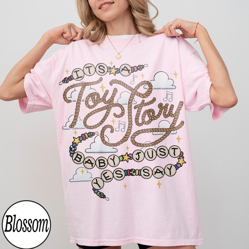May include: A light pink short-sleeve t-shirt featuring a graphic design with the text "ITS A Toy Story BABY JUST SAY YES" written in a rope-style font. The design includes colorful beads, yellow stars, musical notes, and white clouds.