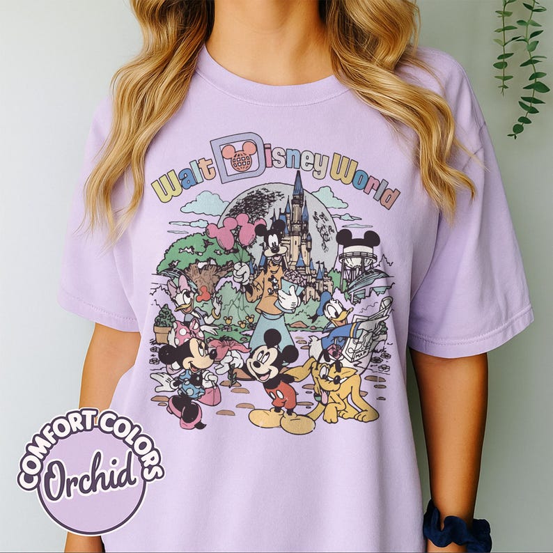 May include: A light purple t-shirt featuring a colorful graphic of Disney characters and the words "Walt Disney World." The design includes Mickey Mouse, Minnie Mouse, Donald Duck, and Goofy in front of a castle. The shirt has the words "Comfort Colors Orchid" on it.