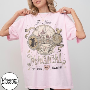 May include: Light pink t-shirt with a graphic design featuring a castle, teacup, and Mickey Mouse. The text on the shirt reads "The Most Magical Place on Earth". The shirt has short sleeves and a round neckline.