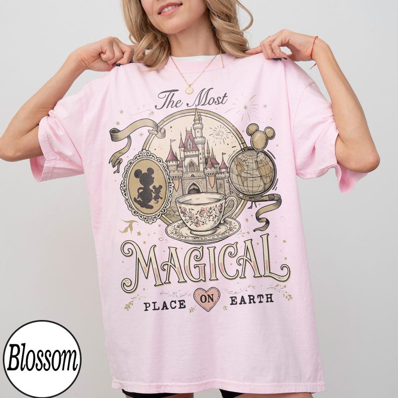 May include: Light pink t-shirt with a graphic design featuring a castle, teacup, and Mickey Mouse. The text on the shirt reads "The Most Magical Place on Earth". The shirt has short sleeves and a round neckline.