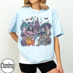 May include: A light blue t-shirt featuring a graphic print of two cartoon characters, one dressed as a pumpkin and the other as a mummy, standing in front of a castle and a moon. The design includes bats, balloons, and a sign reading Hollywood Studios.