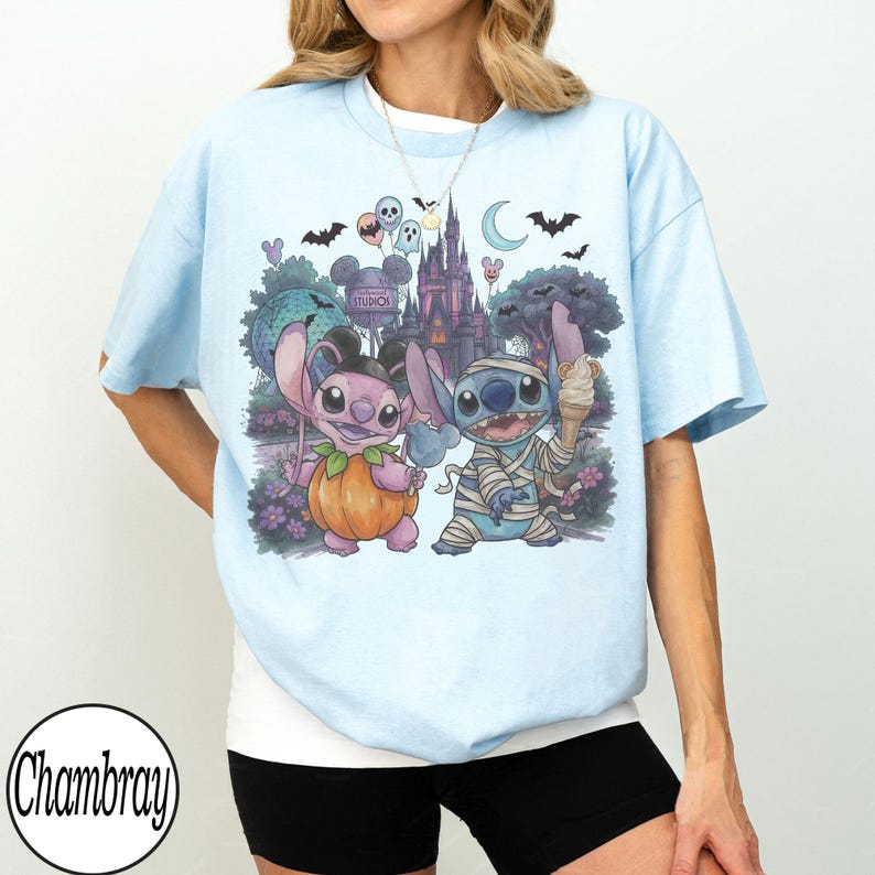 May include: A light blue t-shirt featuring a graphic print of two cartoon characters, one dressed as a pumpkin and the other as a mummy, standing in front of a castle and a moon. The design includes bats, balloons, and a sign reading Hollywood Studios.