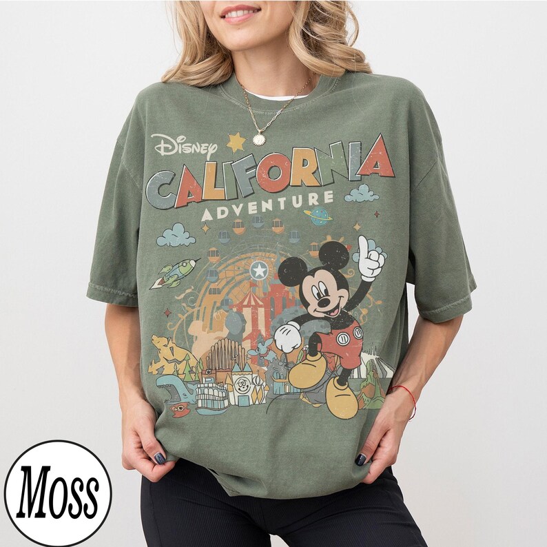 May include: Olive green t-shirt featuring the text "Disney California Adventure" with a graphic of Mickey Mouse. The design includes a Ferris wheel, a rocket, and other amusement park elements. The shirt has short sleeves and a crew neck.