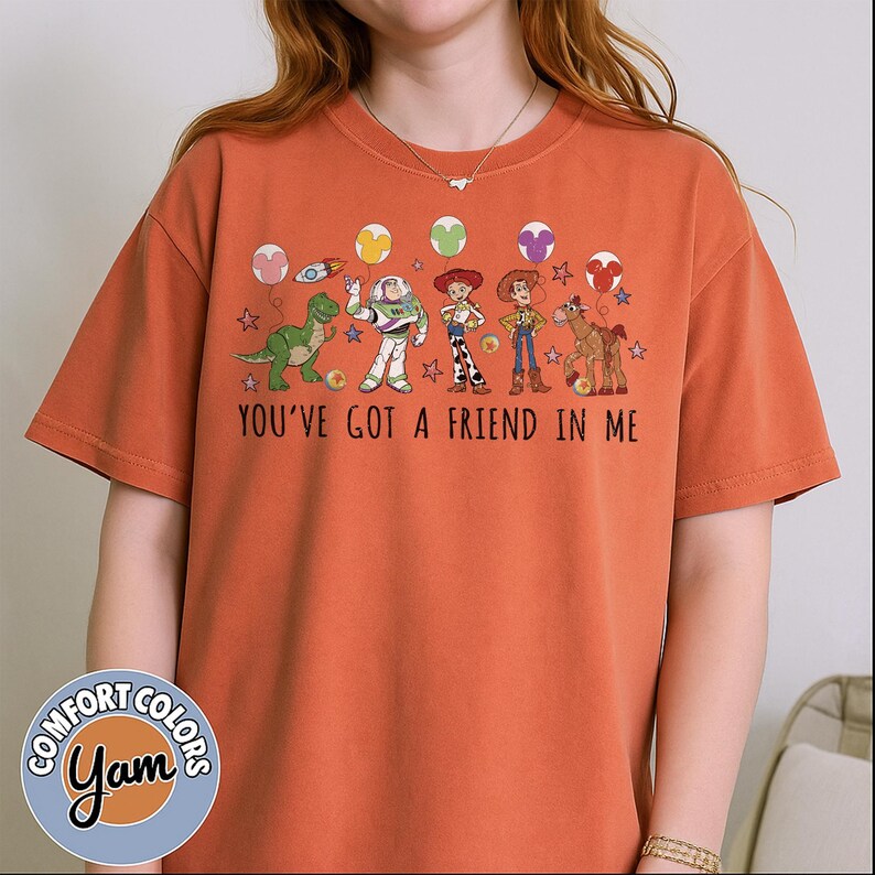 May include: Orange Comfort Colors t-shirt featuring a graphic of Toy Story characters, including Rex, Buzz Lightyear, Woody, and Jessie. The text "You've Got a Friend in Me" is printed below the characters. The shirt has a round neck and short sleeves.