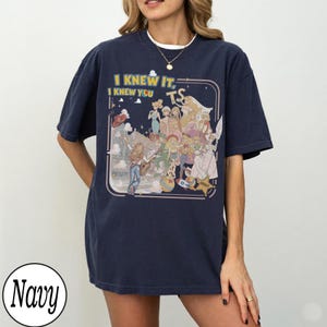 May include: A navy blue short-sleeve t-shirt featuring a graphic print of various animated characters and the text I KNEW IT, I KNEW YOU TS. The design includes a central illustration of multiple figures, a rocket, a ball, and a sheriff star badge.