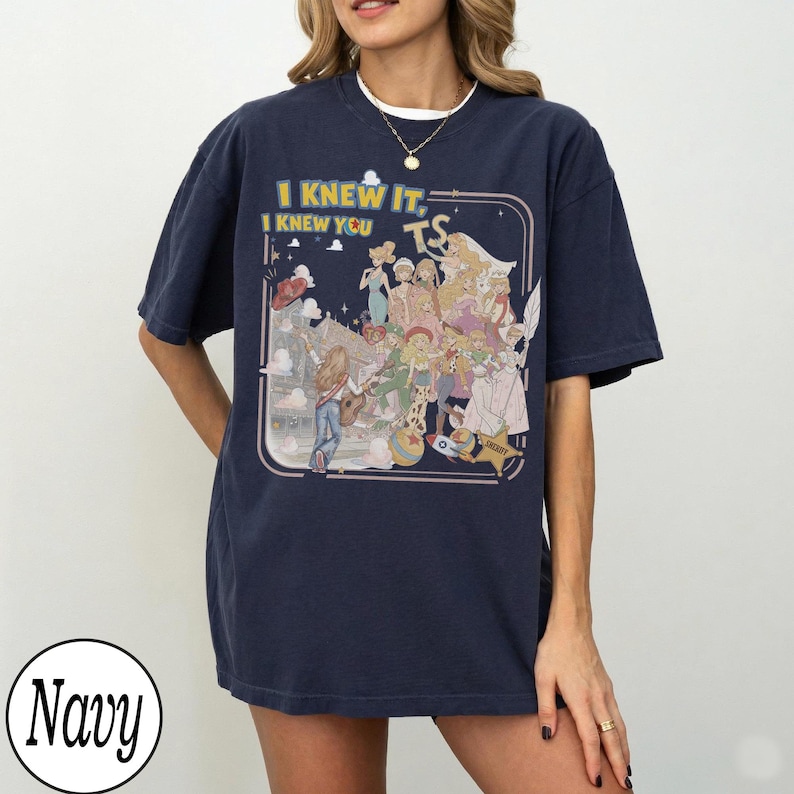 May include: A navy blue short-sleeve t-shirt featuring a graphic print of various animated characters and the text I KNEW IT, I KNEW YOU TS. The design includes a central illustration of multiple figures, a rocket, a ball, and a sheriff star badge.