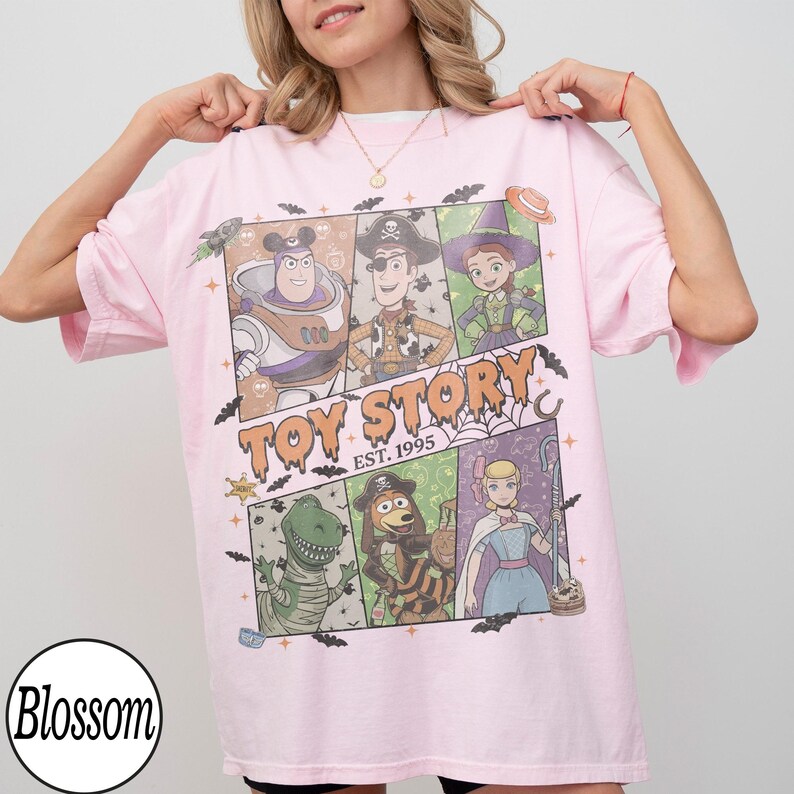 May include: A light pink t-shirt featuring a graphic print of Toy Story characters in Halloween costumes. The design includes Buzz Lightyear, Woody, Jessie, Rex, Slinky Dog, and Bo Peep, with the text Toy Story Est. 1995 and scattered bats.