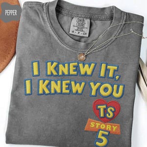 Puede incluir: Una camiseta gris carb&oacute;n con el texto bordado I KNEW IT, I KNEW YOU en amarillo con contornos azules. Debajo del texto hay un coraz&oacute;n rojo que contiene las letras TS, seguido de la palabra STORY y el n&uacute;mero 5 en amarillo y azul.