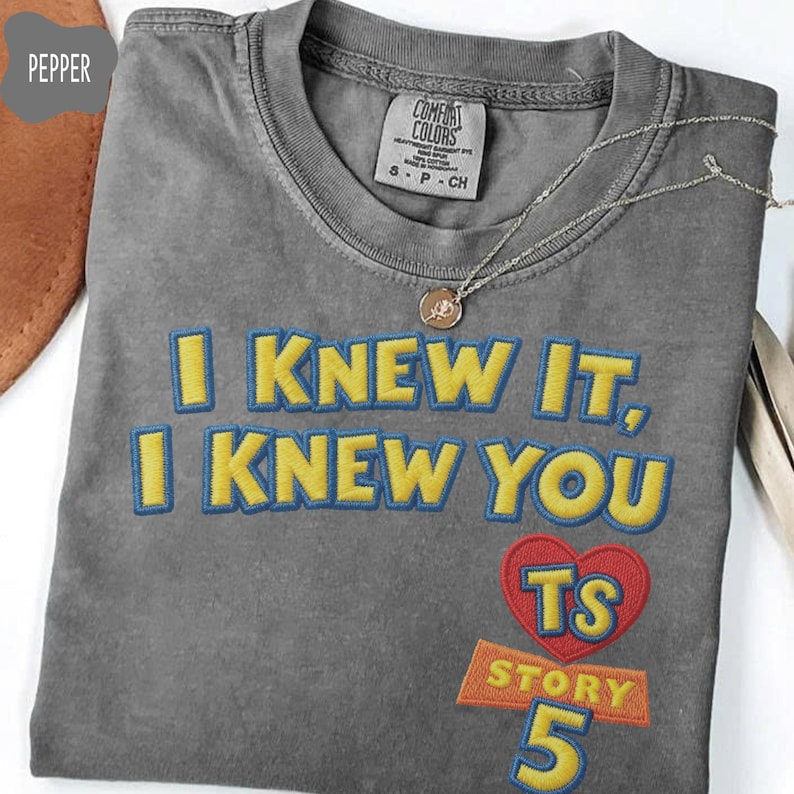Puede incluir: Una camiseta gris carb&oacute;n con el texto bordado I KNEW IT, I KNEW YOU en amarillo con contornos azules. Debajo del texto hay un coraz&oacute;n rojo que contiene las letras TS, seguido de la palabra STORY y el n&uacute;mero 5 en amarillo y azul.