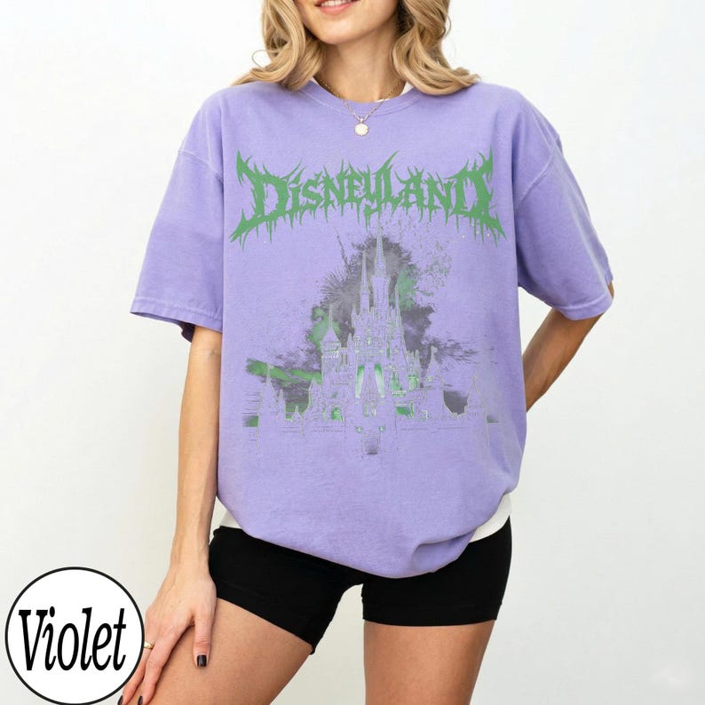 May include: A violet-colored t-shirt with the word "Disneyland" in green, stylized text. The shirt features a graphic of a castle with green accents. The model is wearing black shorts.