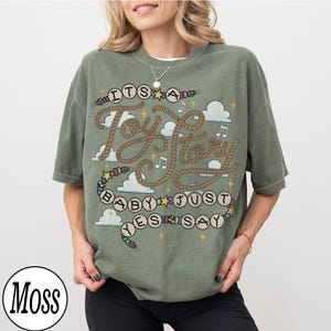 May include: A moss green short-sleeve t-shirt featuring a graphic print with the text "ITS A Toy Story BABY JUST YES SAY" written in rope-style lettering and beads. The design includes white clouds, yellow stars, and musical notes.