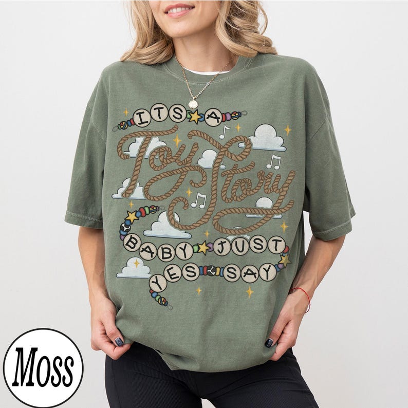 May include: A moss green short-sleeve t-shirt featuring a graphic print with the text "ITS A Toy Story BABY JUST YES SAY" written in rope-style lettering and beads. The design includes white clouds, yellow stars, and musical notes.