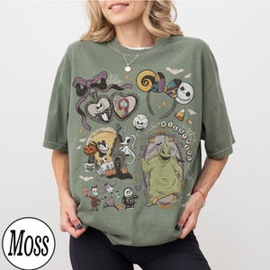 May include: Moss green t-shirt featuring a collage of characters and elements from "The Nightmare Before Christmas." The design includes Jack Skellington, Sally, Zero, Oogie Boogie, and other iconic imagery. The shirt has a relaxed fit.