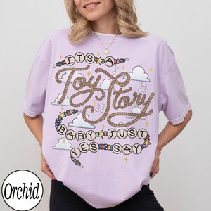 May include: A light purple t-shirt featuring a graphic design with the text "ITS A Toy Story BABY JUST YES SAY". The design includes a rope-style font, colorful beads, stars, musical notes, and white clouds printed on the front of the garment.