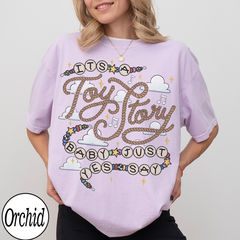 May include: A light purple t-shirt featuring a graphic design with the text "ITS A Toy Story BABY JUST YES SAY". The design includes a rope-style font, colorful beads, stars, musical notes, and white clouds printed on the front of the garment.