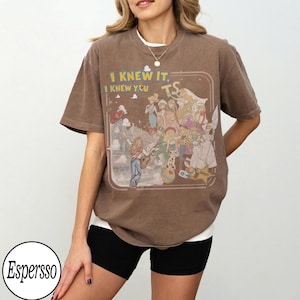 May include: A brown short-sleeve t-shirt featuring a graphic print of various animated characters and the text "I KNEW IT, I KNEW YOU" in yellow and white. The design is enclosed in a rounded rectangular frame with a light-colored background.