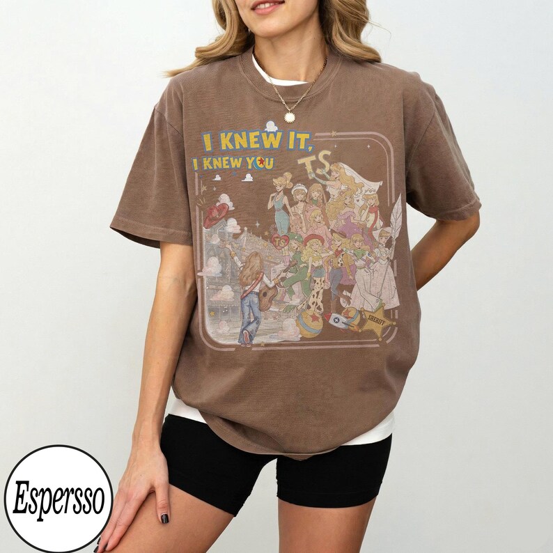 May include: A brown short-sleeve t-shirt featuring a graphic print of various animated characters and the text "I KNEW IT, I KNEW YOU" in yellow and white. The design is enclosed in a rounded rectangular frame with a light-colored background.