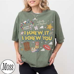 May include: A moss green t-shirt featuring a graphic print with the text I KNEW IT I KNEW YOU, infinity and beyond, and YOU'VE GOT A FRIEND IN ME. The design includes illustrations of cowboy boots, a red hat, alphabet blocks, a star, and a beaded bracelet.
