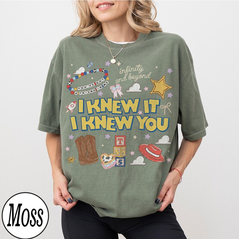 May include: A moss green t-shirt featuring a graphic print with the text I KNEW IT I KNEW YOU, infinity and beyond, and YOU'VE GOT A FRIEND IN ME. The design includes illustrations of cowboy boots, a red hat, alphabet blocks, a star, and a beaded bracelet.