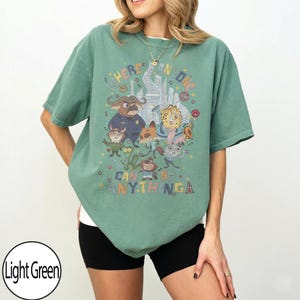 May include: Light green t-shirt with a cartoon graphic and the text "WHERE ANYONE CAN BE ANYTHING". The design features various characters. The shirt has a relaxed fit and short sleeves.