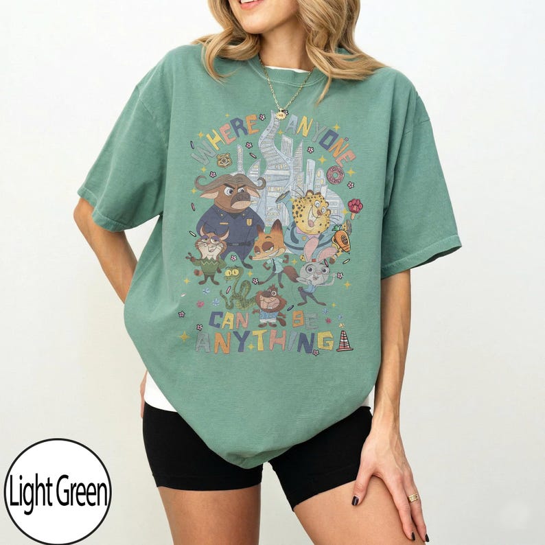 May include: Light green t-shirt with a cartoon graphic and the text "WHERE ANYONE CAN BE ANYTHING". The design features various characters. The shirt has a relaxed fit and short sleeves.