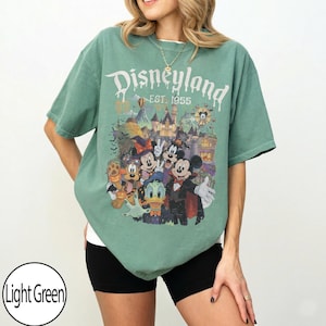 Puede incluir: Una camiseta de manga corta de color verde claro con un estampado gr&aacute;fico de personajes de Disney con disfraces de Halloween. El texto dice Disneyland EST. 1955. El dise&ntilde;o incluye a Mickey Mouse, Minnie Mouse, Donald Duck y Pluto frente a un castillo.