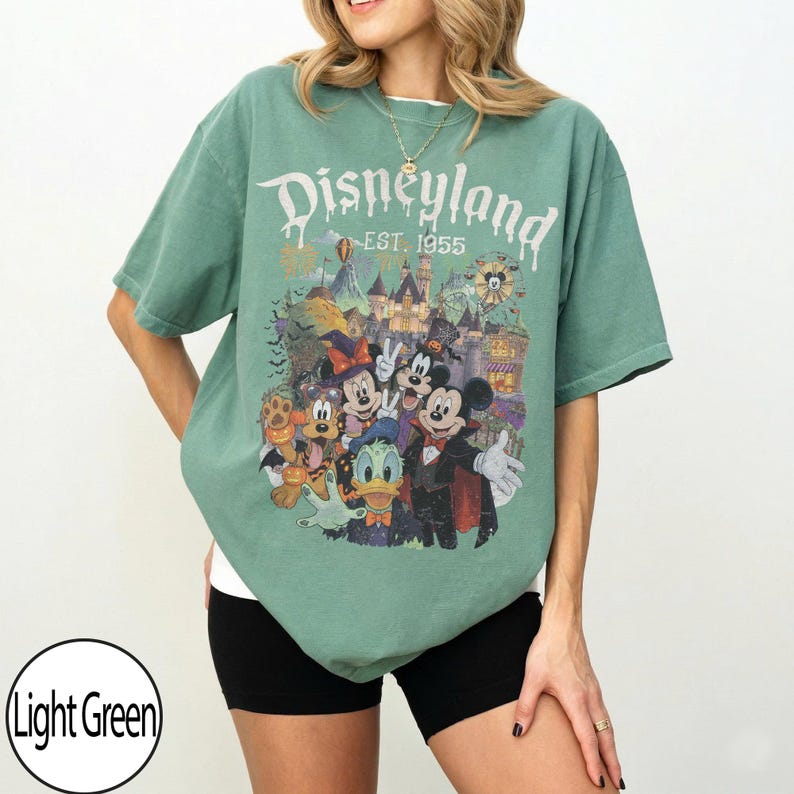 Puede incluir: Una camiseta de manga corta de color verde claro con un estampado gr&aacute;fico de personajes de Disney con disfraces de Halloween. El texto dice Disneyland EST. 1955. El dise&ntilde;o incluye a Mickey Mouse, Minnie Mouse, Donald Duck y Pluto frente a un castillo.