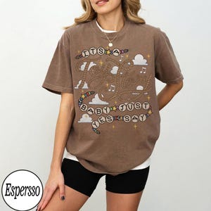 May include: A brown short-sleeve t-shirt featuring a graphic design with rope-style lettering that reads "ITS A Toy Story BABY JUST YES SAY". The design includes small colorful beads, white clouds, musical notes, and yellow stars scattered throughout.