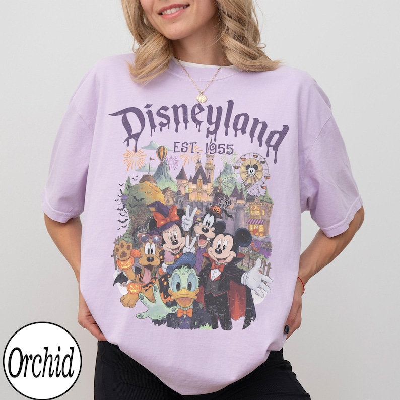 Puede incluir: Una camiseta de manga corta de color morado claro con un estampado gr&aacute;fico de personajes de Disney con disfraces de Halloween frente a un castillo. El texto dice Disneyland EST. 1955. El dise&ntilde;o incluye a Mickey, Minnie, Donald y Pluto.