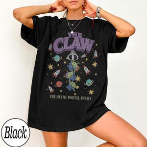 Puede incluir: Una camiseta negra de manga corta con un estampado gr&aacute;fico que incluye el texto THE CLAW y THE MYSTIC PORTAL AWAITS. El dise&ntilde;o muestra tres alien&iacute;genas verdes de tres ojos, una garra mec&aacute;nica, cohetes coloridos, planetas y estrellas.