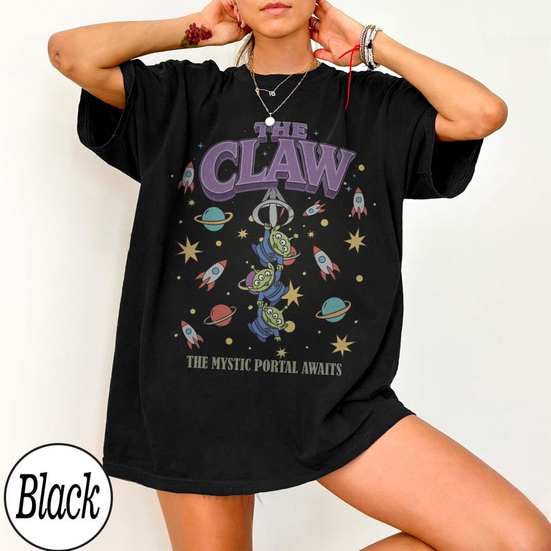 Puede incluir: Una camiseta negra de manga corta con un estampado gr&aacute;fico que incluye el texto THE CLAW y THE MYSTIC PORTAL AWAITS. El dise&ntilde;o muestra tres alien&iacute;genas verdes de tres ojos, una garra mec&aacute;nica, cohetes coloridos, planetas y estrellas.