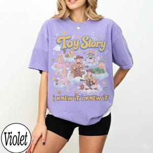 Może przedstawiać: Jasnofioletowy t-shirt z kr&oacute;tkim rękawem z nadrukiem graficznym przedstawiającym postacie z Toy Story w stylu anime. Projekt zawiera napis "Toy Story" u g&oacute;ry oraz "i KNEW iT, i KNEW iT" u dołu, z r&oacute;żnymi postaciami umieszczonymi na chmurach.