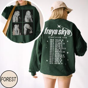 May include: Forest green sweatshirt featuring a black and white image of a singer in four squares. The back of the sweatshirt displays the text "freya skye STARS ALIGN TOUR" and tour dates. A round label with the word "FOREST" is at the bottom.