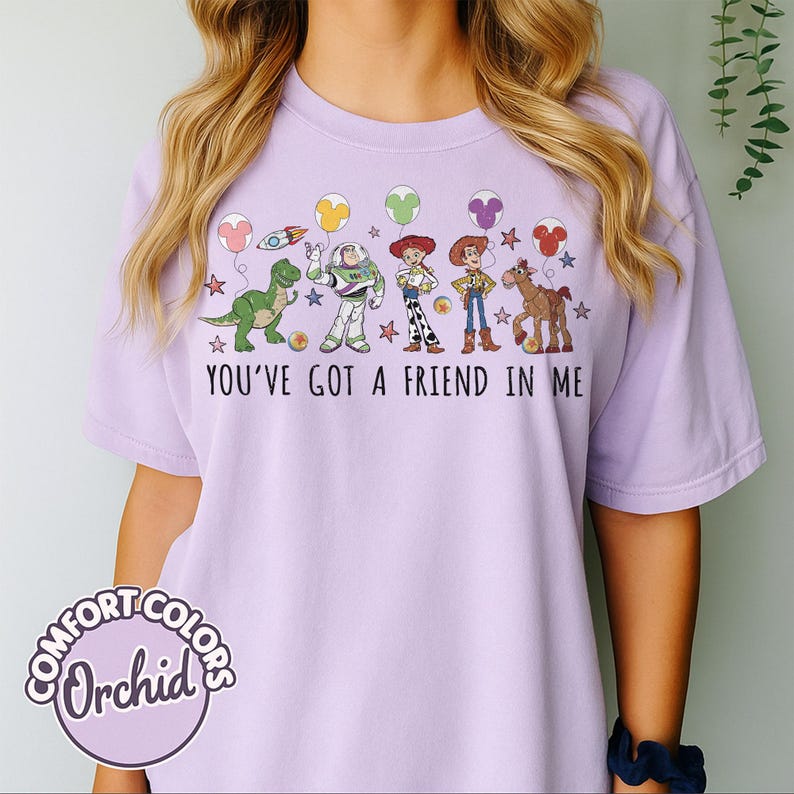 May include: A light purple t-shirt with a graphic featuring cartoon characters and the text "YOU'VE GOT A FRIEND IN ME." The design includes a dinosaur, Buzz Lightyear, Woody, and a horse, with star and balloon accents. The shirt has a Comfort Colors Orchid label.