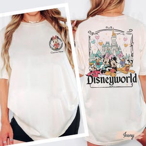 May include: Ivory-colored t-shirt featuring a Minnie Mouse graphic on the front and a Disney World design on the back. The front graphic includes a checkered border and the text "Custom Name." The back graphic showcases Disney characters and the text "Disneyworld 1971."
