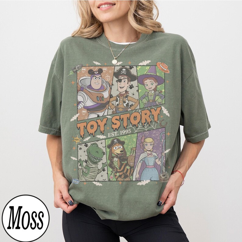 May include: A moss green short-sleeve t-shirt featuring a graphic print of Toy Story characters in Halloween costumes. The design includes Buzz Lightyear, Woody, Jessie, Rex, Slinky Dog, and Bo Peep with the text Toy Story Est. 1995.