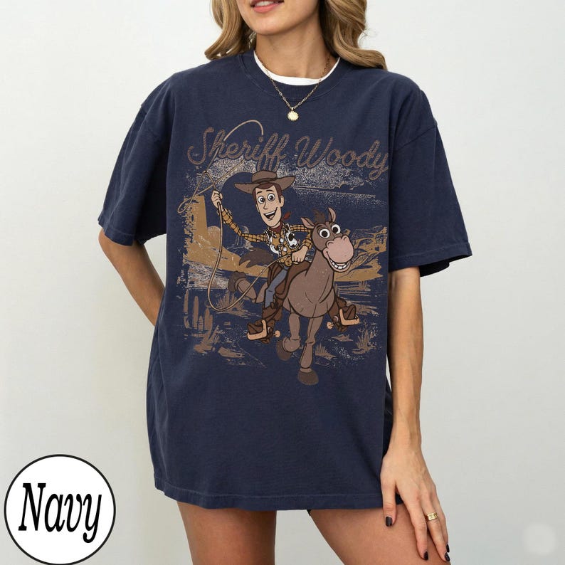 May include: Navy blue t-shirt featuring a graphic of Sheriff Woody riding a horse with the text "Sheriff Woody". The design is in shades of brown and tan, with a vintage aesthetic. The shirt is a relaxed fit.