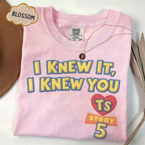 Puede incluir: Una camiseta rosa claro con el texto bordado I KNEW IT, I KNEW YOU en amarillo con bordes azules. Debajo del texto hay un coraz&oacute;n rojo que contiene las letras TS, seguido de la palabra STORY y el n&uacute;mero 5 en amarillo y azul.