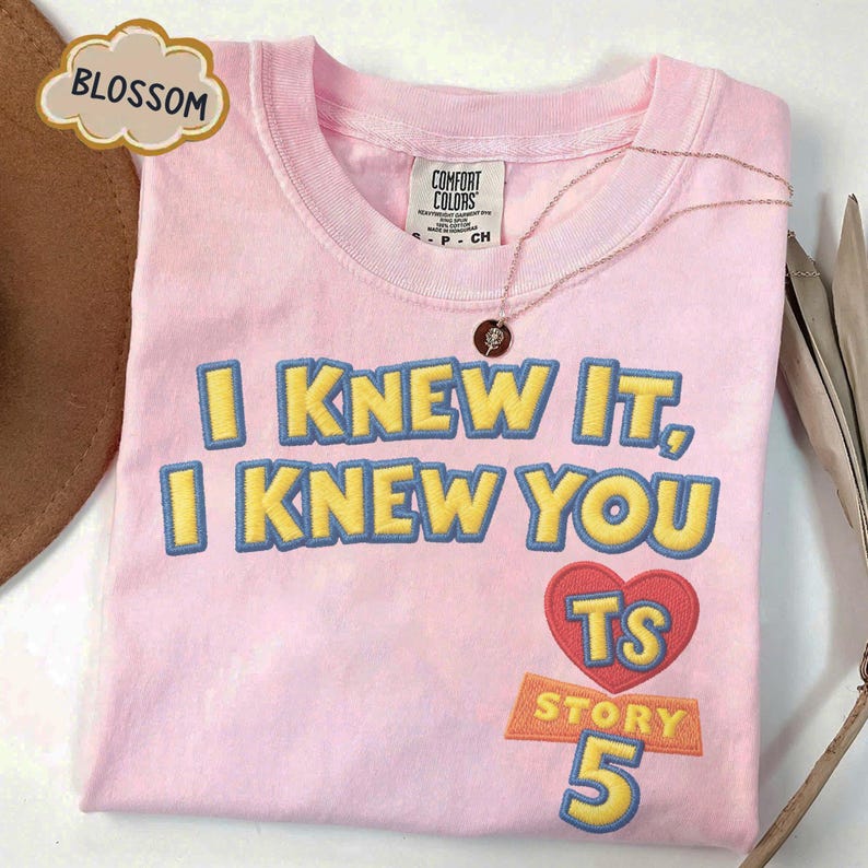 Puede incluir: Una camiseta rosa claro con el texto bordado I KNEW IT, I KNEW YOU en amarillo con bordes azules. Debajo del texto hay un coraz&oacute;n rojo que contiene las letras TS, seguido de la palabra STORY y el n&uacute;mero 5 en amarillo y azul.