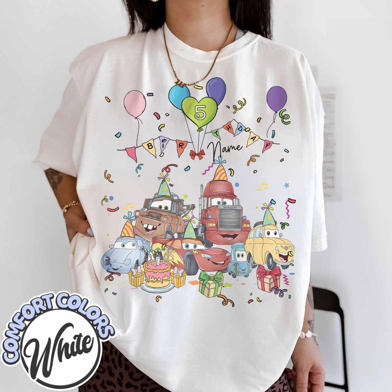 Peut inclure: T-shirt blanc Comfort Colors avec un motif de f&ecirc;te d'anniversaire de voitures de dessins anim&eacute;s color&eacute;s. Le motif comprend des voitures, des ballons, un g&acirc;teau et le chiffre 5. Le mot "Name" est &eacute;galement visible.