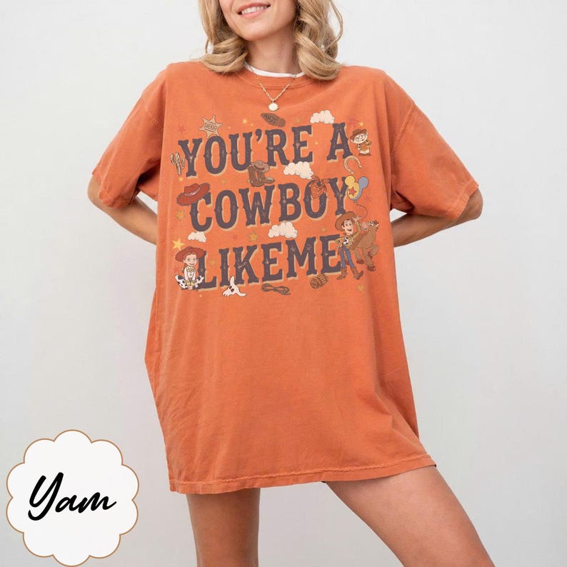 May include: An oversized burnt orange t-shirt featuring the text "YOU'RE A COWBOY LIKEME" in dark blue lettering. The graphic includes illustrations of cowboy boots, hats, a lasso, and cartoon characters in a western style on the front.