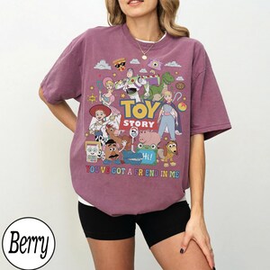 May include: A berry-colored t-shirt featuring a graphic print of Toy Story characters including Woody, Buzz Lightyear, and Jessie. The text reads TOY STORY and YOU'VE GOT A FRIEND IN ME. The design includes various colorful icons and movie figures.