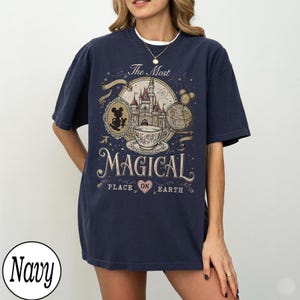 May include: Navy blue t-shirt with a graphic design featuring a castle, teacup, and Mickey Mouse. The text reads "The Most Magical Place on Earth." The design is in shades of beige, brown, and white.
