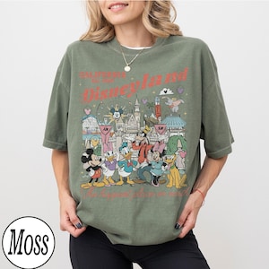 May include: An olive green t-shirt with a Disneyland graphic featuring Mickey Mouse, Minnie Mouse, Donald Duck, Goofy, and Pluto. The text "California Est. 1955 Disneyland" is printed in red. The shirt has a vintage, distressed appearance.