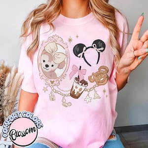 Könnte beinhalten: Rosa Comfort Colors T-Shirt mit Cartoon-Design. Das Design umfasst einen gerahmten Bären, Mickey Mouse Ohren, ein Getränk und eine Brezel. Goldene Kettenakzente und das Wort "Blossom" in einem Kreis sind ebenfalls enthalten.