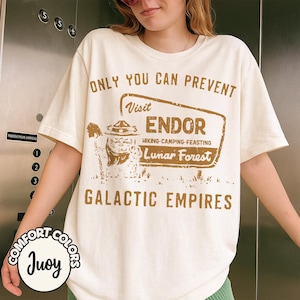 Puede incluir: Camiseta color crema con un gráfico y texto marrón. El gráfico presenta un personaje y las palabras "Visit Endor, Lunar Forest". El texto en la camiseta dice "Only You Can Prevent Galactic Empires."