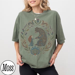 以下が含まれることがあります： オリーブグリーンのTシャツ。円形のデザインで、クマと女の子が手をつないでおり、花や動物のイラストで囲まれています。デザインは茶色、青、緑の色合いを使用しています。Tシャツは半袖で、ゆったりとしたフィット感です。